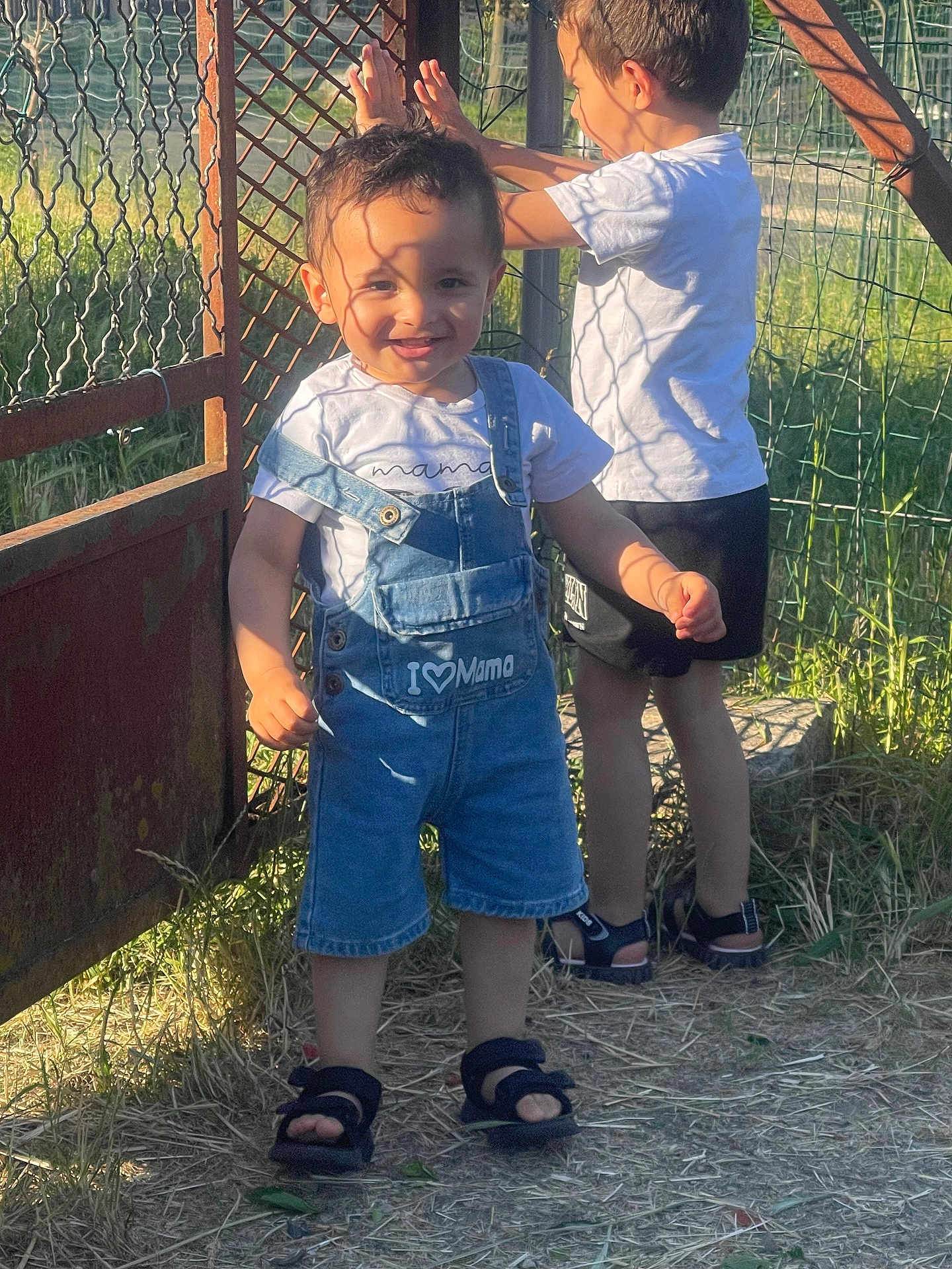 Iyaas a rejoint le concours — aidez-le/la à gagner de superbes lots ! child, boy, smiling, outdoor, sunlight, shadow, denim_overalls, tshirt, sandals, grass, metal_gate, fence, nature, play, summer, happy, two_children, portrait, daylight, casual_clothing