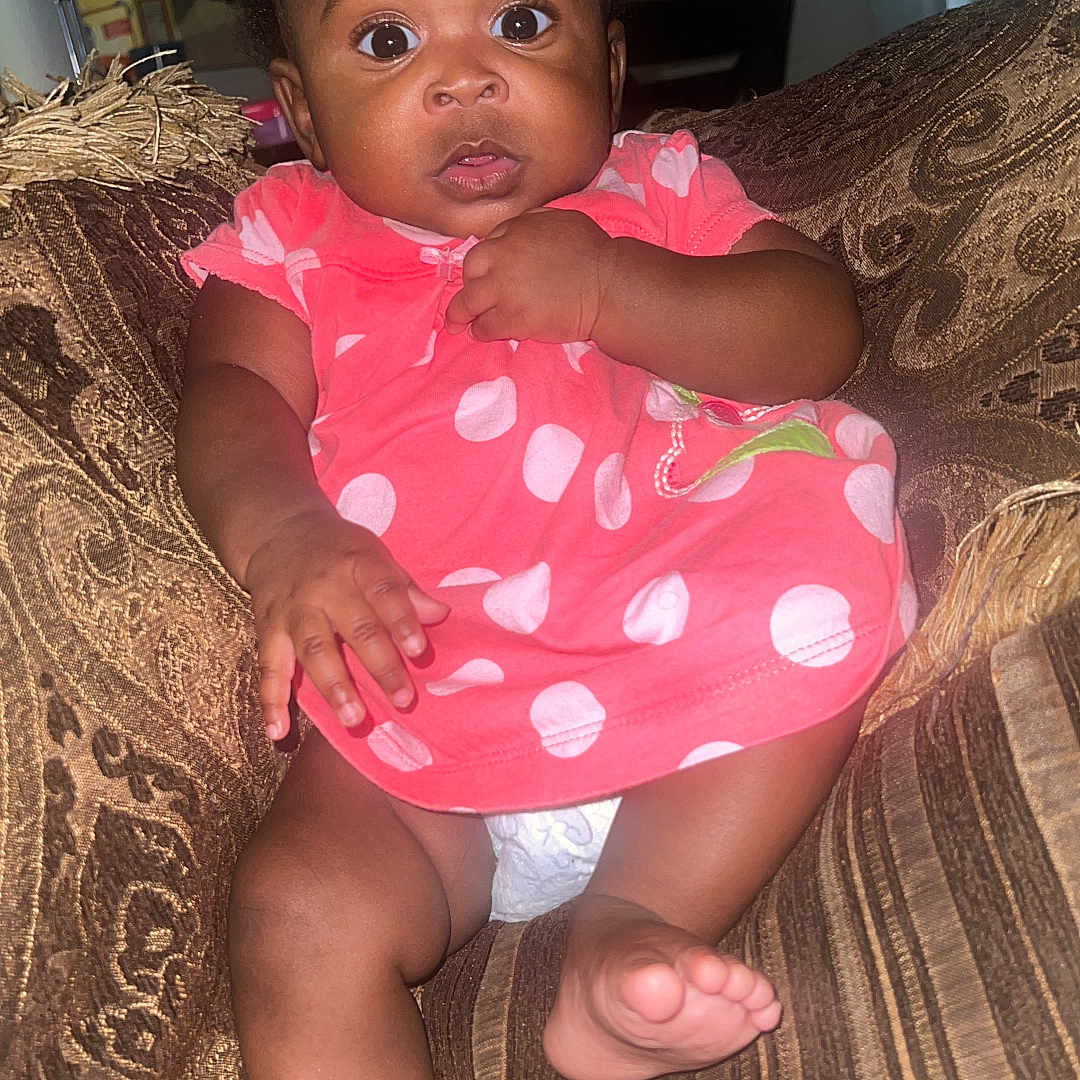 Eliayne Cacho joined the competition — help win amazing prizes! baby, child, infant, dress, pink, polka_dot, couch, sofa, brown, patterned, furniture, indoor, foot, hand, curious, wide_eyes, person, seated, home, cute