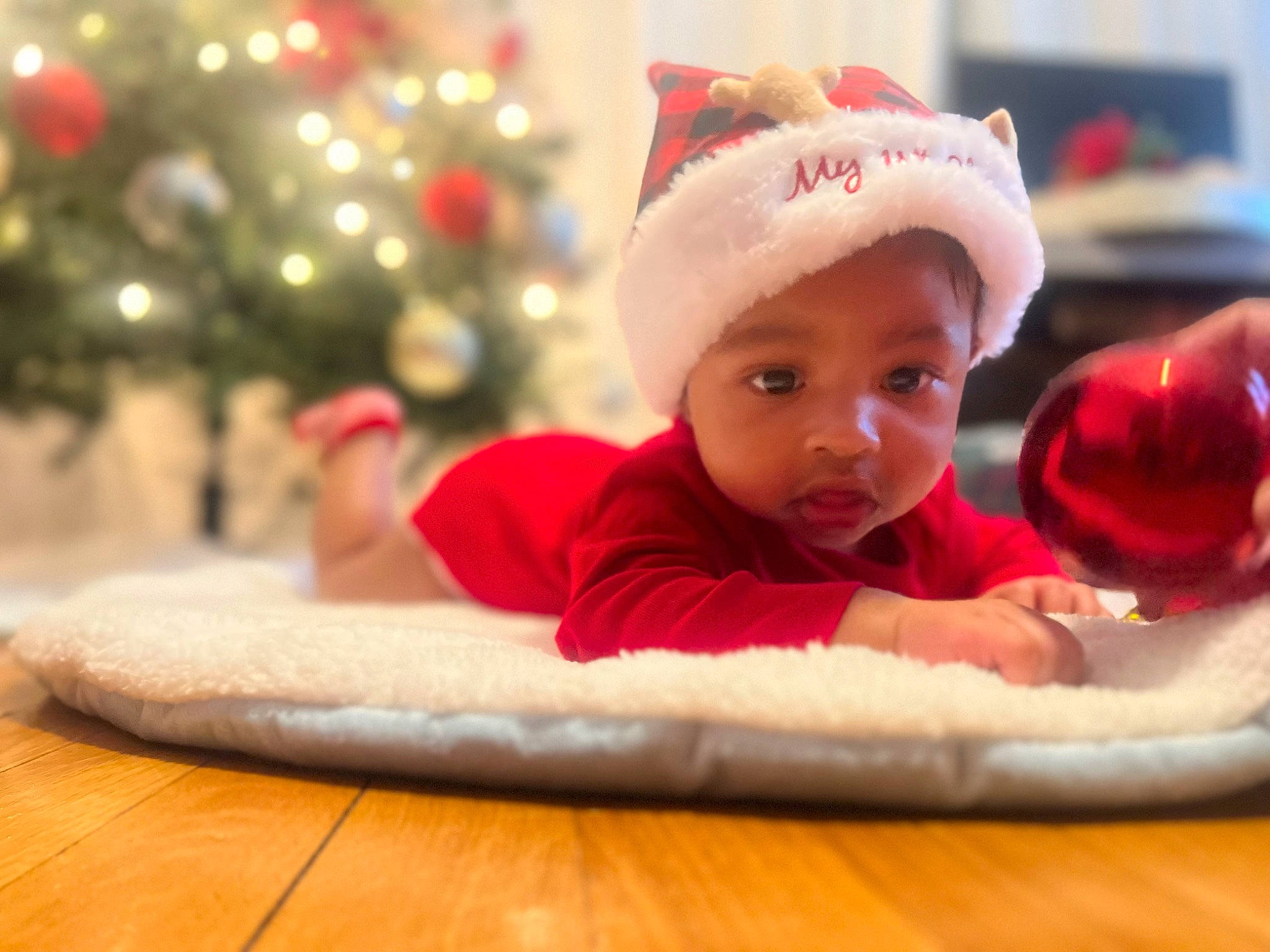 Ezra De Carvalho is registered to the contest to win money with this photo: baby, baby_toddler_clothing, cap, carmine, child, christmas, christmas_eve, christmas_tree, comfort, event, fictional_character, flooring, fun, happy, hardwood, headwear, holiday, person, room, santa_claus