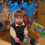 élias participe au concours pour gagner de l'argent avec cette photo : toddler, child, book, reading, christmas_tree, nutcracker, teddy_bear, holiday_decorations, rug, toy_puzzle, indoor, festive, winter, decorations, clothing, socks, carpet, wooden_furniture, face, portrait