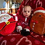 baby, plush_toy, santa_pillow, gingerbread_pillow, red_blanket, heart_pattern, peppermint_pattern, indoor, child, holiday_theme, christmas, soft_toys, furniture, lamp, wooden_chair, table, window_light, cozy, cute, portrait