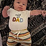Braxlee is registered to the contest to win money with this photo: baby, infant, child, clothing, shirt, shorts, striped_shorts, patterned_blanket, lying_down, person, cute, young_child, smiling, arm_raised, indoors, text_on_clothing, baby_face, soft_lighting, casual, relaxed