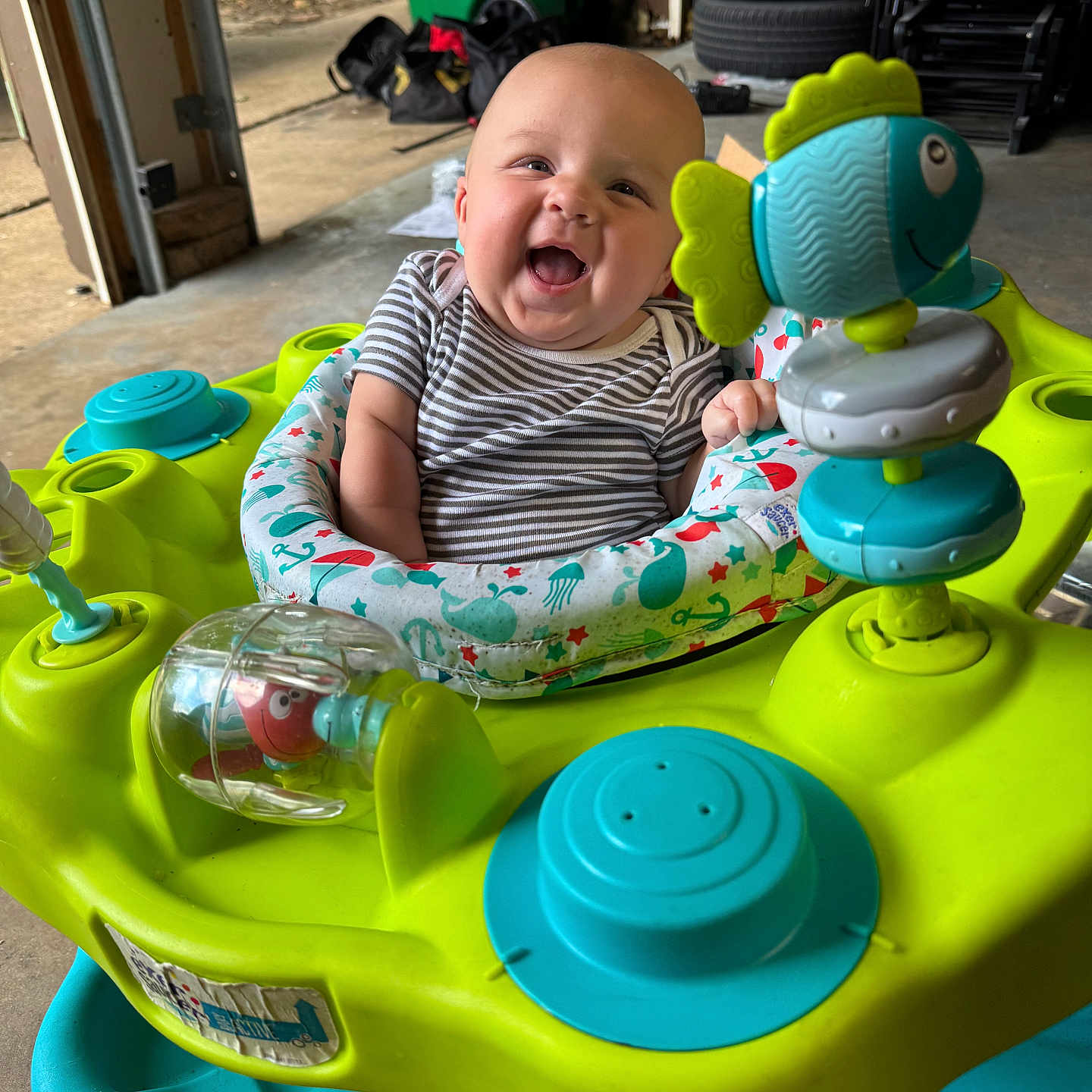 Braxlee is registered to the contest to win money with this photo: activity_center, baby, bag, child, clothing, colorful, expression, floor, garage, happy, indoor, infant, person, plastic, playful, seat, smiling, striped_shirt, tire, toy