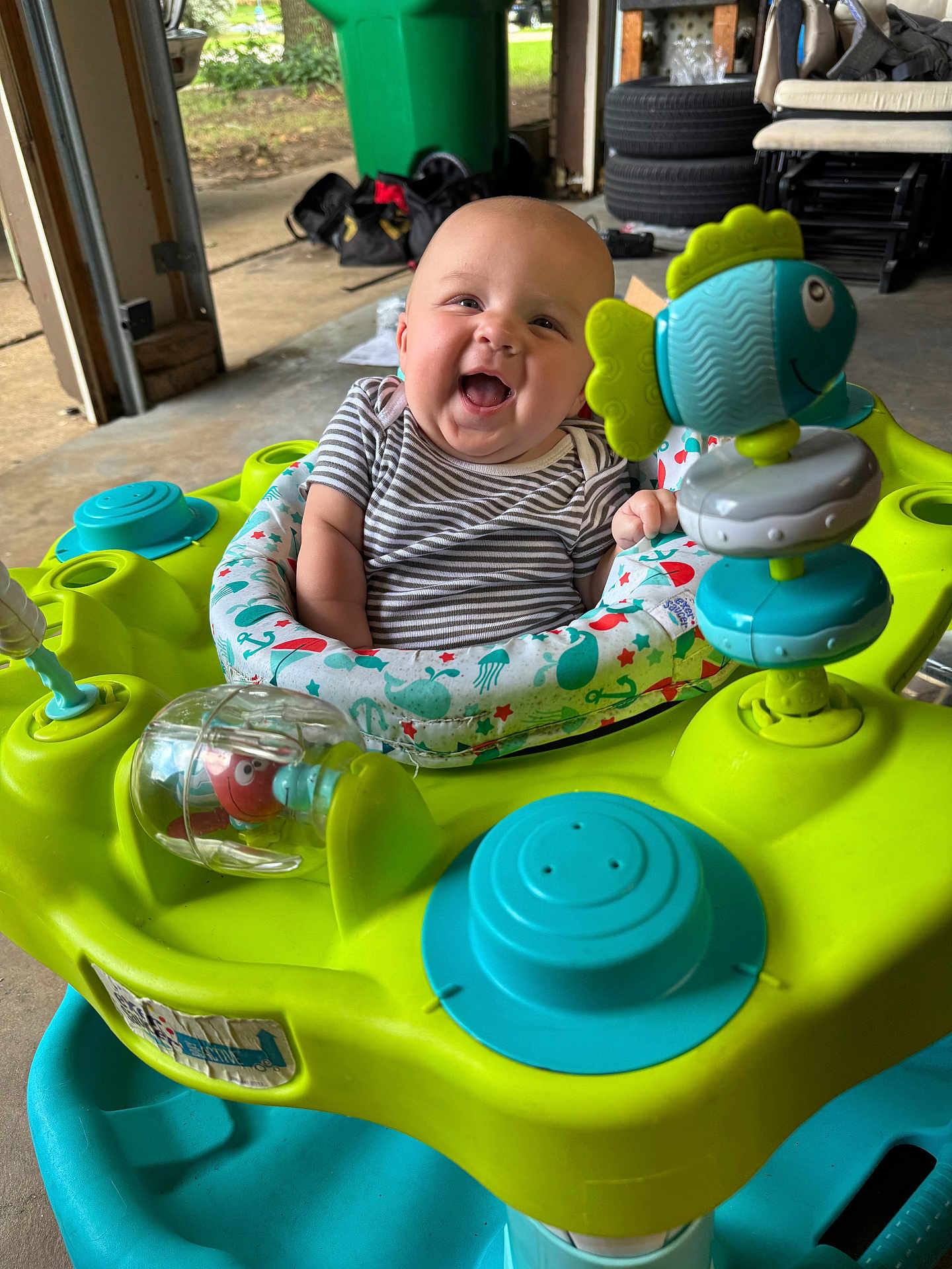 Braxlee is registered to the contest to win money with this photo: baby, infant, smiling, activity_center, toy, indoor, child, playful, happy, person, seat, colorful, clothing, striped_shirt, plastic, garage, tire, bag, floor, expression