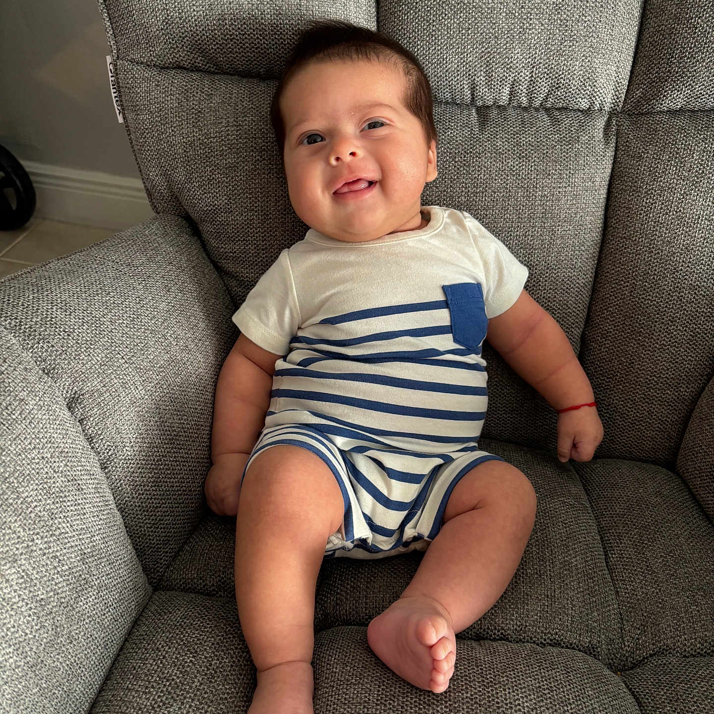 Jordan is registered to the contest to win money with this photo: armchair, baby, child, cozy, cute, face, foot, furniture, gray_fabric, hand, happy, indoors, infant, onesie, portrait, red_bracelet, sitting, skin, smiling, striped_clothing