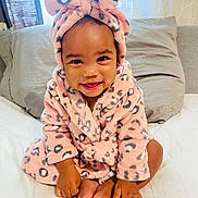 Milli Lorraine is registered to the contest to win money with this photo: baby, child, smiling, pink, robe, headband, bow, leopard_print, bed, pillow, curtain, indoors, cute, happy, infant, person, skin, foot, hand, sitting