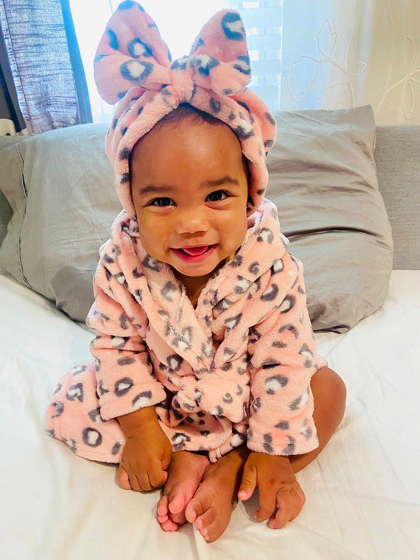 Milli Lorraine is registered to the contest to win money with this photo: baby, child, smiling, pink, robe, headband, bow, leopard_print, bed, pillow, curtain, indoors, cute, happy, infant, person, skin, foot, hand, sitting