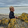 child, boy, smiling, rocks, lichen, coast, seaside, cloudy_sky, outdoor, nature, water, shore, landscape, casual_clothing, happy, playful, daylight, scenic, youth, relaxing