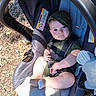 baby, child, car_seat, outdoor, sunlight, blue_eyes, hooded_clothing, socks, wood_chips, seatbelt, infant, cute, smiling, person, closeup, portrait, playground, shade, fabric, toy
