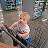 Michael is registered to the contest to win money with this photo: baby, shopping_cart, store, child, blond_hair, blue_eyes, toddler, hand, grocery_store, aisle, shelf, products, floor, clothing, person, shopping, holding, cute, indoor, curious