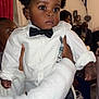 baby, child, bow_tie, white_shirt, curly_hair, indoor, person, hand, fuzzy_sleeve, seated_people, curious_expression, family_gathering, casual_clothing, black_pants, pacifier_clip, decor, curtains, portrait, closeup, holding