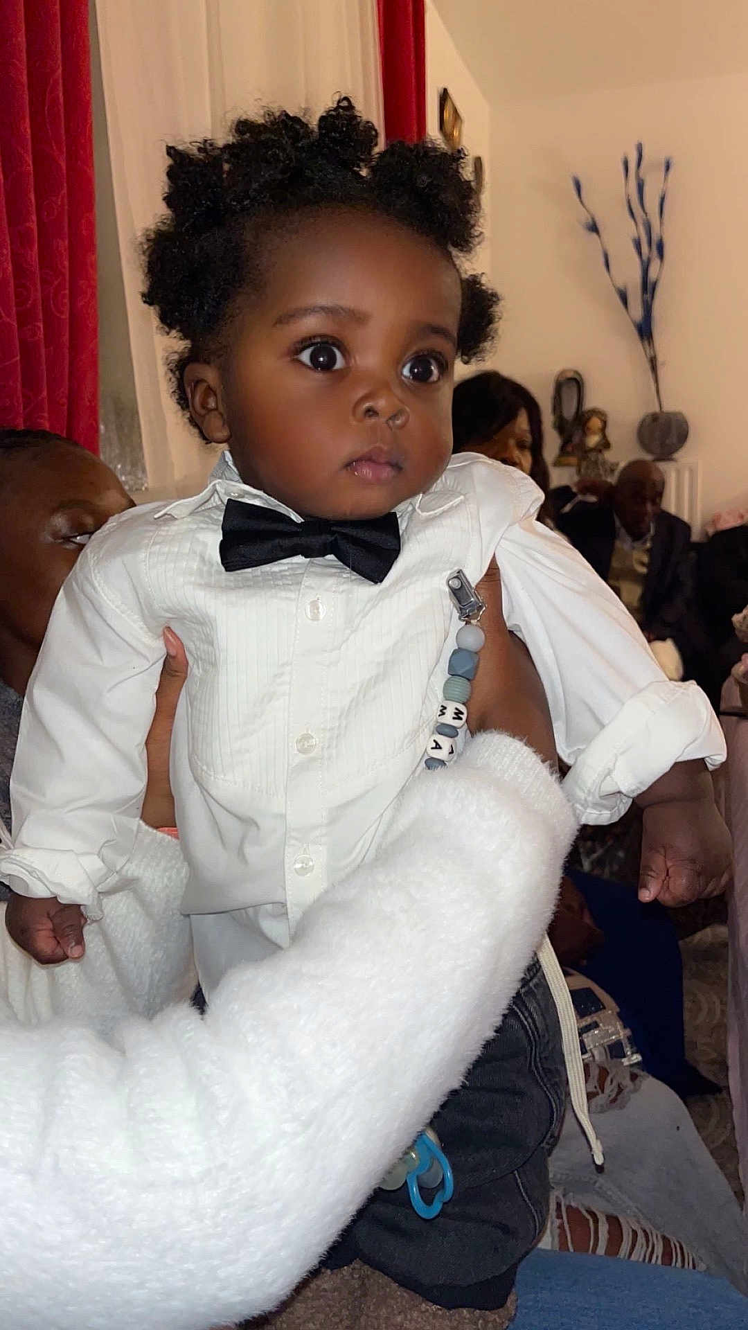 Mayron participe au concours pour gagner de l'argent avec cette photo : baby, child, bow_tie, white_shirt, curly_hair, indoor, person, hand, fuzzy_sleeve, seated_people, curious_expression, family_gathering, casual_clothing, black_pants, pacifier_clip, decor, curtains, portrait, closeup, holding