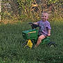 Sky joined the competition — help win amazing prizes! baby, child, toy, tractor, green, yellow, grass, outdoor, fence, chain_link_fence, plants, purple_shirt, person, cute, sitting, play, sunlight, nature, yard, summer