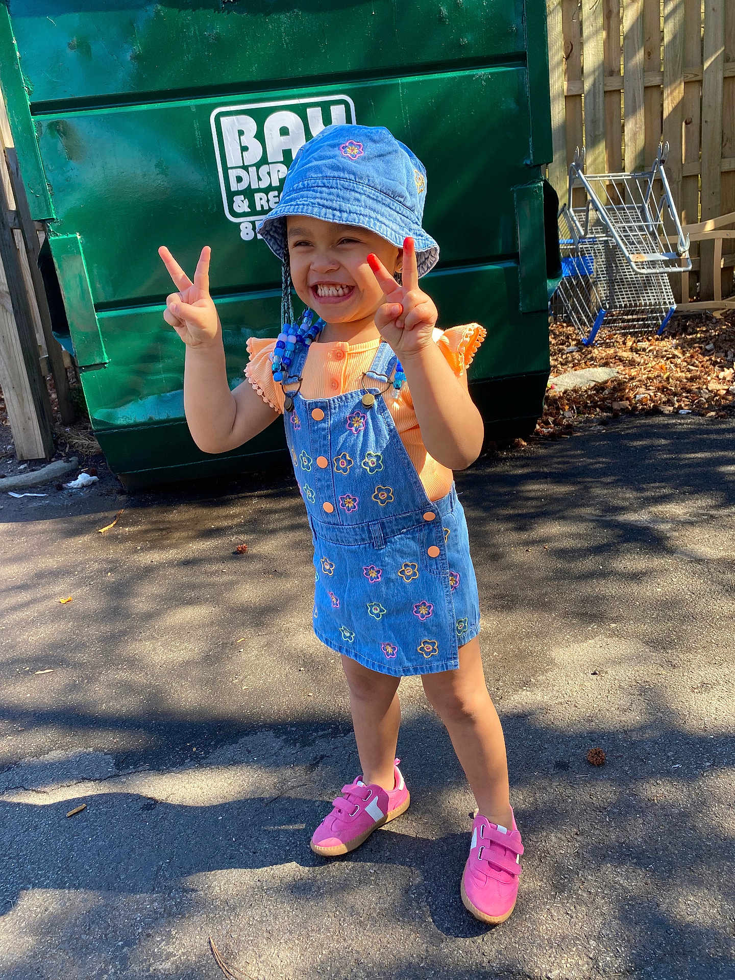 Bloom joined the competition — help win amazing prizes! child, girl, smiling, peace_sign, bucket_hat, denim_dress, orange_shirt, pink_shoes, outdoor, pavement, sunlight, shopping_cart, dumpster, fence, beads, happy, fashion, casual, playful, portrait