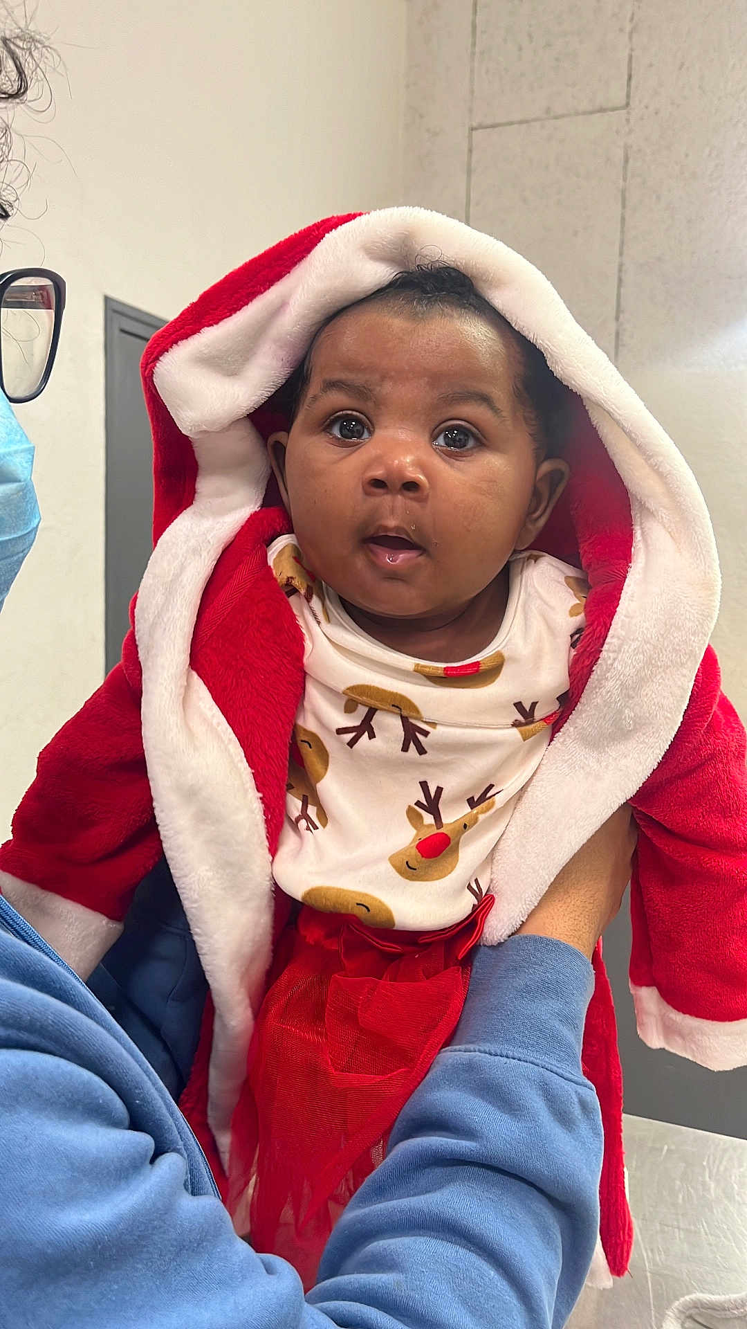 Anna Joëlle participe au concours pour gagner de l'argent avec cette photo : baby, child, person, face, hood, red_clothing, white_fur, reindeer_print, festive, holiday, indoor, curious, blue_sweatshirt, mask, hands, closeup, portrait, cute, warm_clothing, expression