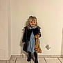 child, girl, blonde_hair, coat, blue_dress, blanket, pacifier, smile, indoor, tile_floor, curly_hair, boots, standing, wall, happy, person, portrait, young_child, casual_clothing, accessory