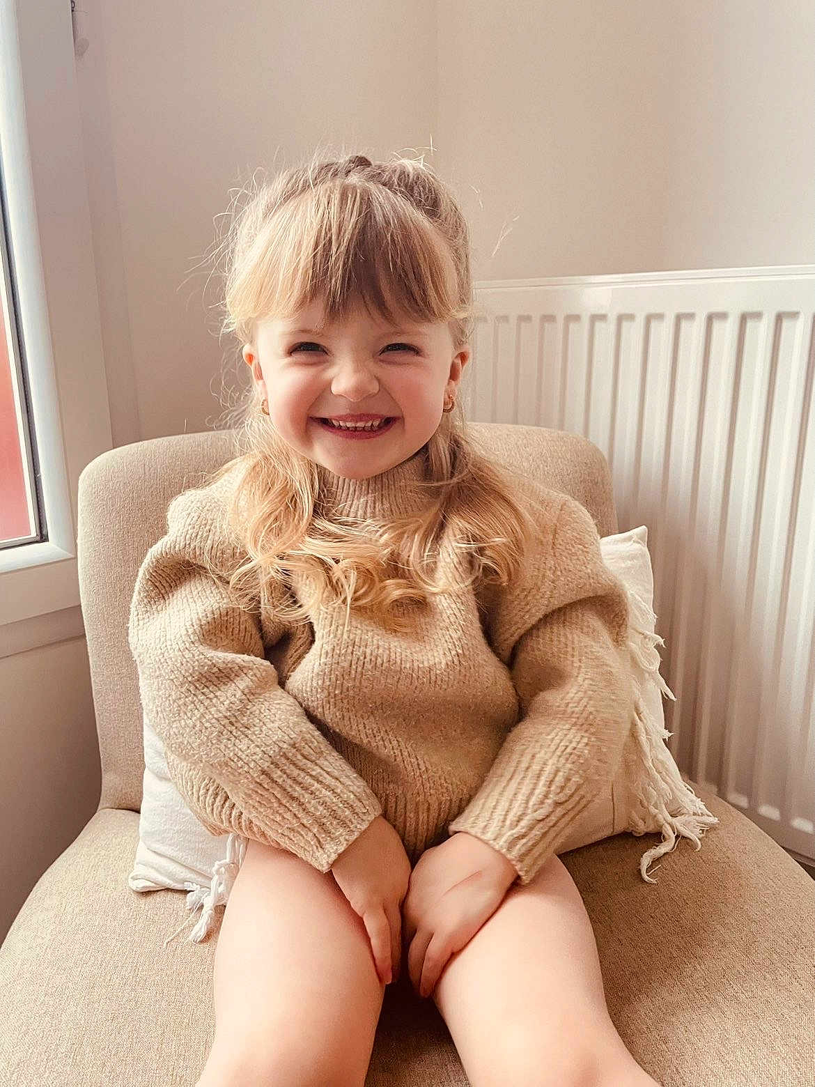 Ambre a rejoint le concours — aidez-le/la à gagner de superbes lots ! child, smiling, girl, sweater, blonde_hair, chair, indoor, cozy, happy, portrait, window, natural_light, curtain, radiator, cushion, casual_clothing, seated, cute, young, warm