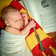 Charlie joined the competition — help win amazing prizes! baby, newborn, sleeping, blanket, scarf, red, yellow, wizard_wand, book, open_book, text, wood_floor, hand, face, portrait, cozy, magic, child, indoor, peaceful