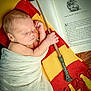 baby, newborn, sleeping, blanket, scarf, red, yellow, wizard_wand, book, open_book, text, wood_floor, hand, face, portrait, cozy, magic, child, indoor, peaceful