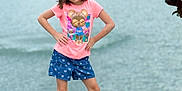 Lilly joined the competition — help win amazing prizes! child, girl, posing, standing, lake, water, grass, outdoor, nature, pink_tshirt, cartoon_print, blue_shorts, white_shoes, smiling, confident, summer, casual_clothing, greenery, young, playful