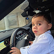 Albert joined the competition — help win amazing prizes! baby, child, toddler, car, driver, steering_wheel, dashboard, vehicle, portrait, indoors, sitting, white_shirt, long_sleeves, short_hair, wide_eyes, looking_at_camera, hands, automotive, road_trip, cute