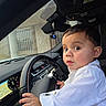 baby, child, toddler, car, driver, steering_wheel, dashboard, vehicle, portrait, indoors, sitting, white_shirt, long_sleeves, short_hair, wide_eyes, looking_at_camera, hands, automotive, road_trip, cute
