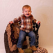 Maverick joined the competition — help win amazing prizes! toddler, child, plaid_shirt, jeans, boots, chair, pumpkin, decor, quilt, smiling, portrait, indoors, fall_theme, neutral_background, cute, happy, seated, fabric, footwear, baby