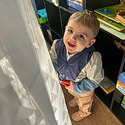 Mayron a rejoint le concours — aidez-le/la à gagner de superbes lots ! bookshelf, child, curtain, face, indoors, jacket, looking_up, natural_light, pants, person, playroom, puzzle, shelf, shoes, smile, smiling, toddler, toy, window, wooden_floor