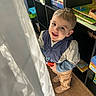 bookshelf, child, curtain, face, indoors, jacket, looking_up, natural_light, pants, person, playroom, puzzle, shelf, shoes, smile, smiling, toddler, toy, window, wooden_floor