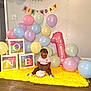 baby, birthday, cake, balloons, colorful, blocks, yellow_rug, indoor, celebration, child, hair_beads, pastel_colors, party, smiling, sitting, floor, number_one_balloon, happy, decorations, cute