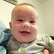 Tyler is registered to the contest to win money with this photo: baby, infant, child, smiling, laughing, portrait, close_up, face, indoors, home, held, hands, clothing, patterned_outfit, gray_shirt, yellow, green, wooden_sign, happy, family