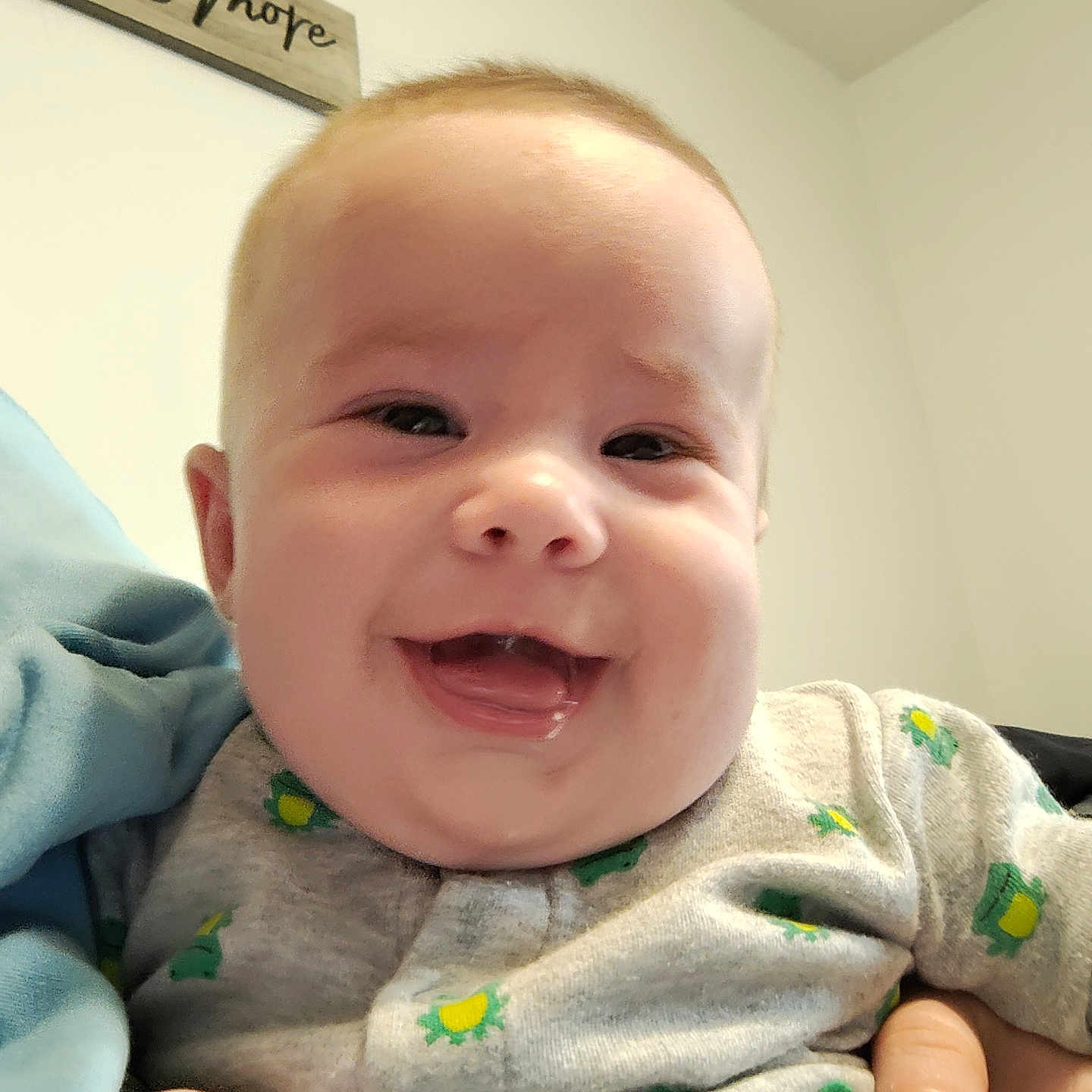 Tyler is registered to the contest to win money with this photo: baby, infant, child, smiling, laughing, portrait, close_up, face, indoors, home, held, hands, clothing, patterned_outfit, gray_shirt, yellow, green, wooden_sign, happy, family