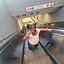 child, girl, escalator, peace_sign, braided_hair, glasses, subway_station, people, stairs, green_shoes, white_shirt, denim_skirt, handrail, public_transport, urban, smile, casual_clothing, indoor, portrait, happy
