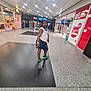 child, shopping_mall, walking, green_slippers, denim_skirt, white_shirt, glasses, indoor, storefront, advertisement, floor, people, lighting, reflection, corridor, retail, shopping, casual, curious, back_view