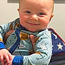 baby, child, smiling, onesie, infant, hand, indoor, person, face, blue_clothing, patterned_clothing, striped_pants, framed_picture, flag, american_flag, patriotic, wall, adult_hand, holding, portrait