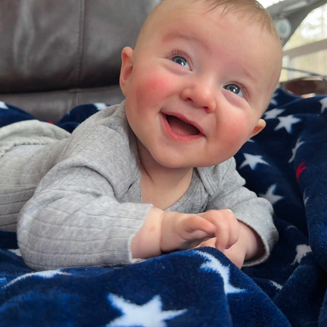 Silas joined the competition — help win amazing prizes! baby, smiling, blanket, star_pattern, blue, indoor, furniture, happy, child, infant, portrait, cozy, clothing, toddler, face, person, head, cute, expression, lying_down