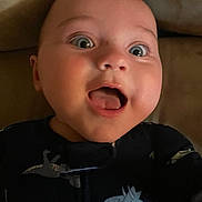 Jameson is registered to the contest to win money with this photo: baby, infant, face, expression, mouth_open, wide_eyes, dinosaur_print, clothing, dark_outfit, close_up, portrait, child, cute, indoor, soft_surface, person, young_child, happy, curious, sleepwear