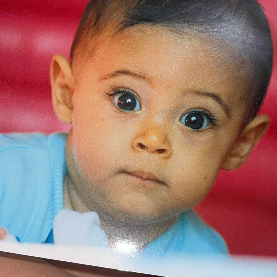 Isleym a rejoint le concours — aidez-le/la à gagner de superbes lots ! arm, baby, black_clothing, blue_clothing, child, closeup, curious, expression, eyes, face, head, holding, indoor, person, photograph, portrait, print, red_background, skin, young_child