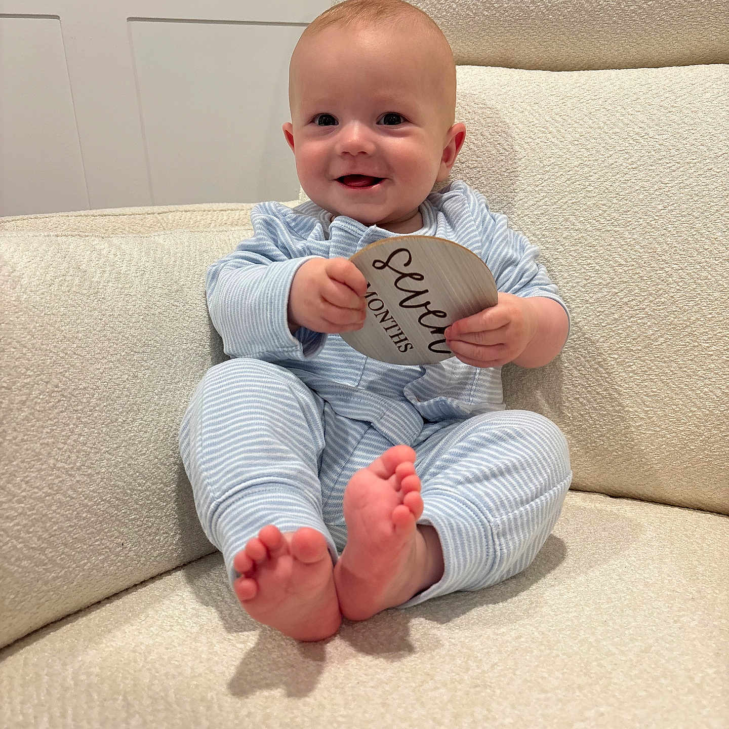 Jackson is registered to the contest to win money with this photo: baby, infant, smiling, pajamas, striped_clothing, couch, furniture, milestone_sign, wooden_sign, seven_months, happy, sitting, barefoot, indoors, soft_texture, child, person, cute, portrait, cozy