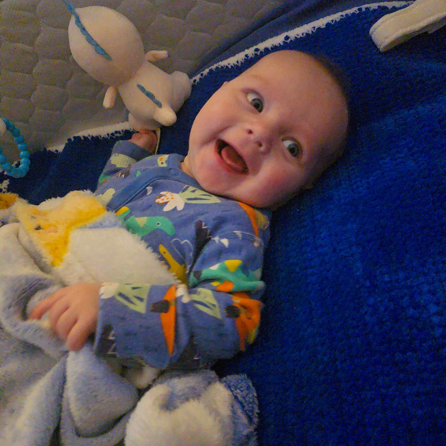 Elias is registered to the contest to win money with this photo: baby, blanket, blue, child, comfort, cozy, cuddly, cute, dinosaur_pajamas, face, happy, indoors, infant, lying_down, plush_toy, sleep_area, smiling, soft, toy, young_child