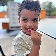 Thyam participe au concours pour gagner de l'argent avec cette photo : child, boy, curly_hair, smiling, outdoor, table, hand, face, eyes, casual_clothing, seat, daylight, cute, portrait, happy, skin, person, sitting, background_blur, young