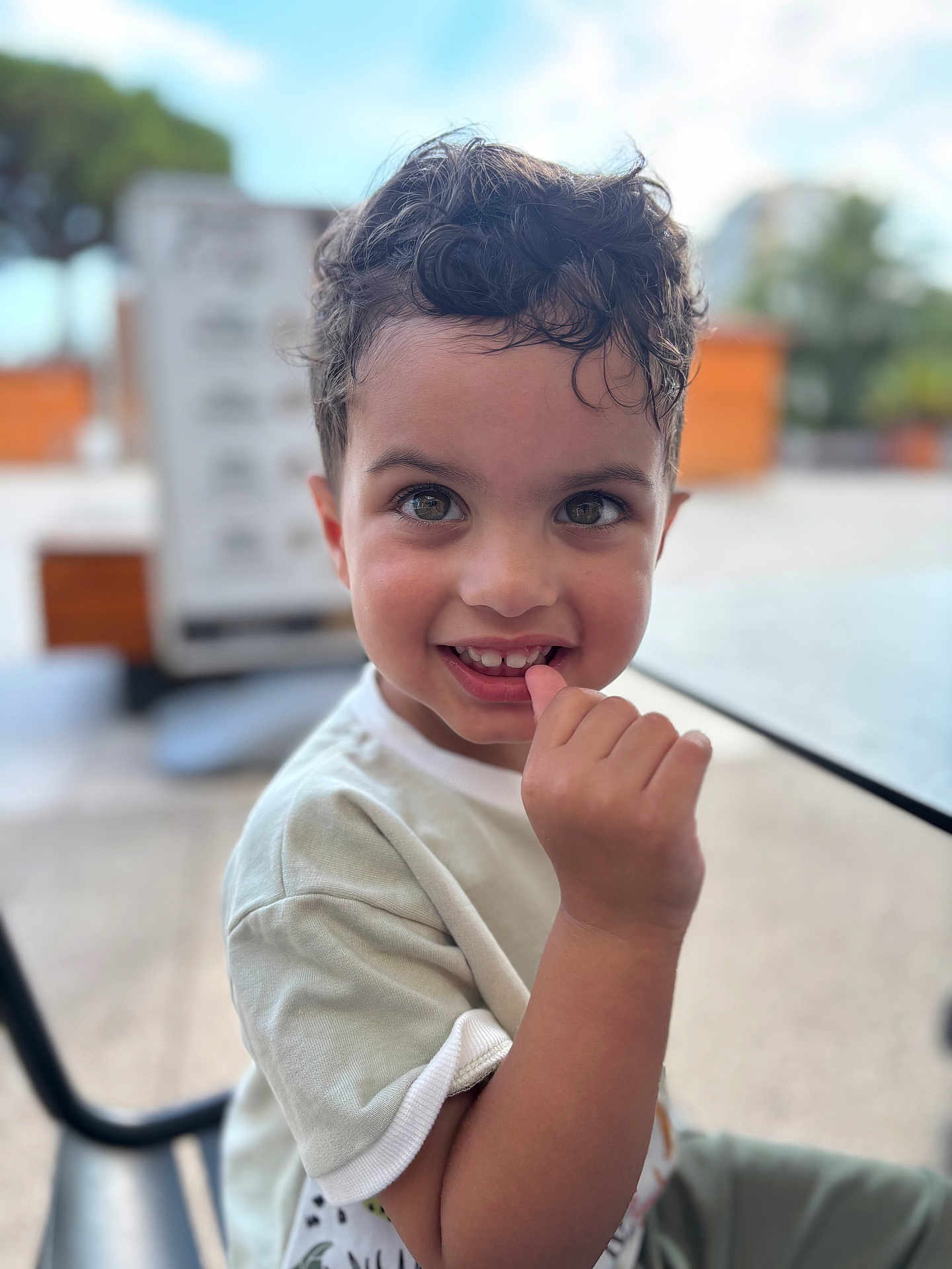 Thyam participe au concours pour gagner de l'argent avec cette photo : child, boy, curly_hair, smiling, outdoor, table, hand, face, eyes, casual_clothing, seat, daylight, cute, portrait, happy, skin, person, sitting, background_blur, young