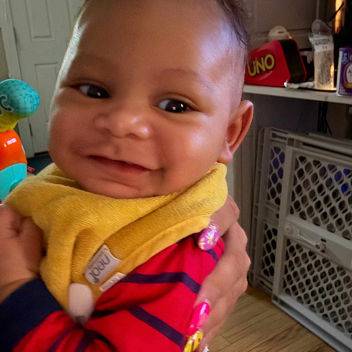 Maison is registered to the contest to win money with this photo: baby, smile, child, indoor, bib, striped_clothing, hand, toy, furniture, door, floor, person, happy, closeup, face, skin, nail_polish, holding, home, portrait