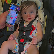 Aurora is registered to the contest to win money with this photo: baby, car_seat, child, infant, phone, hand, toy, colorful, safety_harness, clothing, indoor, cute, portrait, expression, seat, pacifier_clip, face, person, looking, closeup