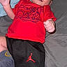 Joseph is registered to the contest to win money with this photo: baby, infant, red_shirt, black_shorts, socks, bed, blanket, smile, happy, face, person, cute, child, indoor, portrait, clothing, young, lying_down, skin, expression