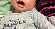 Joseph is registered to the contest to win money with this photo: baby, smiling, onesie, text_on_clothing, bed, blanket, purple_blanket, green_blanket, infant, happy, face, lying_down, cute, person, child, indoors, cozy, soft_texture, mustache_graphic, clothing