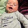 baby, smiling, onesie, text_on_clothing, bed, blanket, purple_blanket, green_blanket, infant, happy, face, lying_down, cute, person, child, indoors, cozy, soft_texture, mustache_graphic, clothing