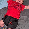 baby, smiling, red_shirt, black_shorts, socks, smiley_face, bed, bedding, happy, infant, child, person, lying_down, indoors, casual_clothing, cute, small, joyful, raised_arms, head