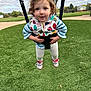 casual_clothing, child, curly_hair, daylight, fun, grass, happy, holding, jacket, nature, outdoor, park, person, play, playground, shoes, smile, swing, toddler, white_leggings
