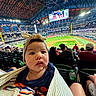 baby, child, stadium, baseball, sports, crowd, lights, hat, headband, blanket, person, seated, field, outdoor, spectator, game, audience, event, daylight, people